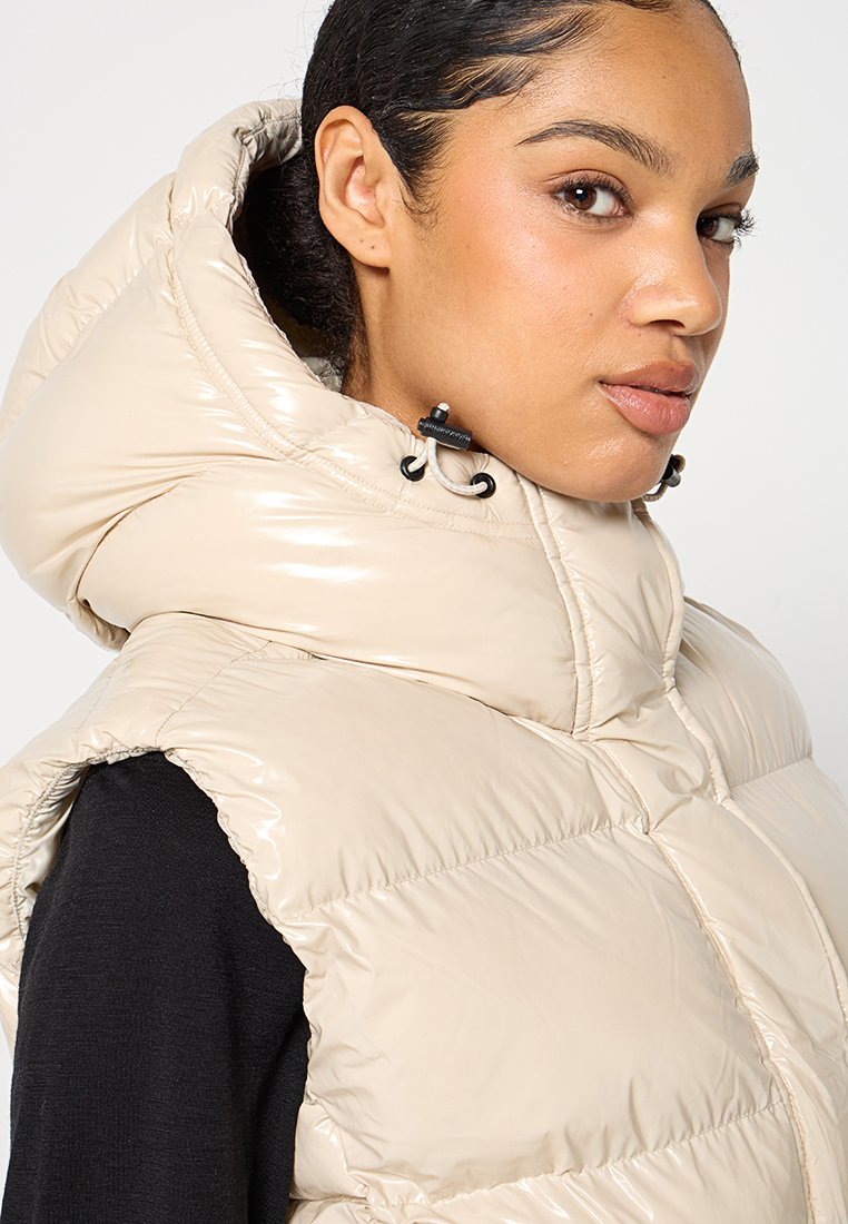 Beige Steppweste mit hohem Kragen, glänzender Oberfläche und sichtbaren Nähten. Verfügt über Toggle-Verschlüsse an der Kapuze. Schwarzes Shirt darunter.