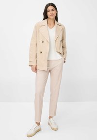 Beige Doppelreiher-Trenchcoat über einem weißen V-Ausschnitt-Oberteil, kombiniert mit hellrosa karierten Hosen und weißen Sneakers mit Jutesohle.