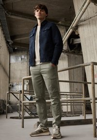 Marineblauwe spijkerjasje, lichtbeige trui, olijfgroene cargobroek met zakken en grijze sneakers met witte veters, geplaatst in een industriële ruimte.