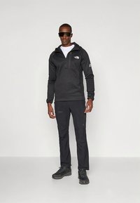 Pulôver de lã negra com meio fecho, com capuz e bolso no peito. Combinado com calças pretas e botas resistentes de caminhada pretas.