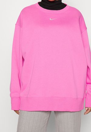 Henkilö, joka pukeutunut vaaleanpunaiseen Nike-huppariin mustan poolopaidan ja vaaleanharmaiden raitahousujen päällä, seisoo yksiväristä taustaa vasten.