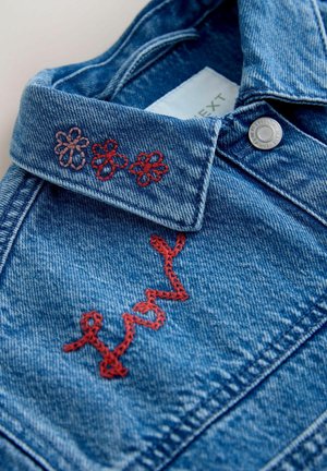 Sinisen farkkutakin kaulus, jossa punaiset kirjotut kukat ja punainen kaunokirjoitus "lovely" hopeanvärisen nepparin lähellä.