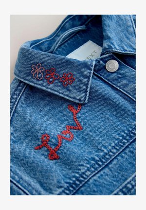 Modrý golier rifľovej bundy s červenými vyšívanými kvetmi a červeným kurzívnym nápisom „lovely“ pri striebornej cvokovej gombíku.