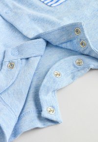 Prenda de algodón azul claro con textura suave, que incluye botones a presión y un detalle de tela a rayas. Costuras visibles y detalles de botones.