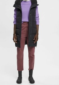 Czarna pikowana kamizelka na zamek i guziki, noszona na teksturowanym fioletowym swetrze, w połączeniu z bordowymi spodniami i czarnymi botkami za kostkę.