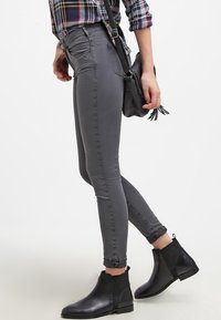 Jeans ajustados grises con una textura suave, cintura de altura media y diseño hasta el tobillo. Combinados con botas negras hasta el tobillo y una camisa de cuadros.