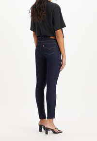 Vaqueros ajustados de color azul oscuro, hechos de denim, con una cintura alta, bolsillos traseros con un logo, combinados con una camiseta corta negra y sandalias de tacón negras.