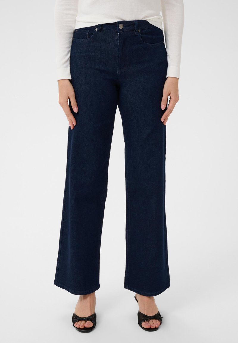 Femme portant un jean large taille haute bleu foncé, un haut blanc à manches longues et des talons ouverts noirs avec des nœuds, debout devant un fond blanc.