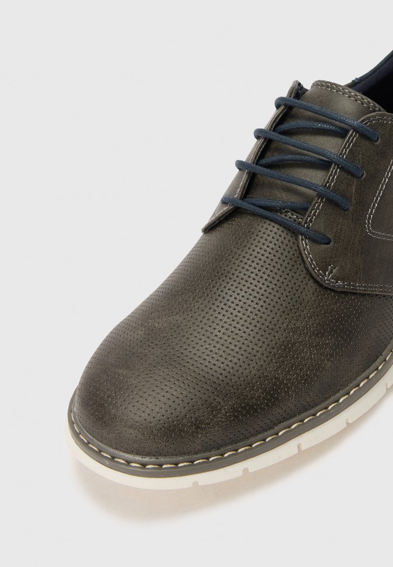 Zapato de cuero gris con superficie texturizada, puntera redonda, cordones en navy y suela de goma blanca. Presenta un patrón perforado a lo largo de la parte superior.