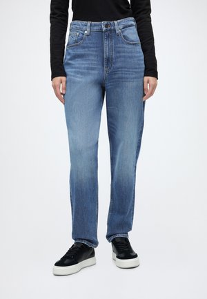 Person iført lyseblå jeans med lige ben, let falmet foran, og sorte sneakers med hvide såler på en hvid baggrund.