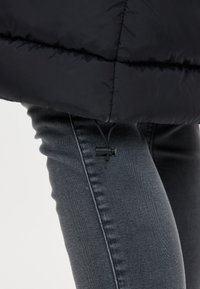 Svart puffjacka med en glänsande yta, med en dragsko vid fållen. Bärs över grå jeans med en slät, strukturerad yta.