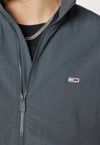Chaqueta gris con cremallera, fabricada en un tejido ligero, que cuenta con cierre de cremallera, cuello alto y un pequeño logo bordado en el pecho.