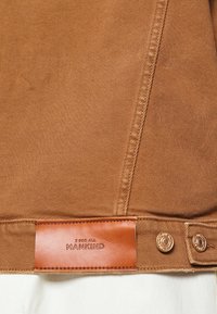 Tissu en denim marron avec une texture lisse, comportant une étiquette en cuir embossée avec "7 FOR ALL MANKIND" et deux boutons en métal.