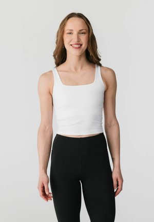 Ung kvinde smilende, iført en hvid ærmeløs crop top og sorte højttaljede leggings, stående mod en ensartet lys baggrund.