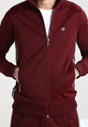 Hombre con chaqueta burdeos con cremallera, manos en los bolsillos, camiseta blanca visible y tatuajes en el cuello y las muñecas.