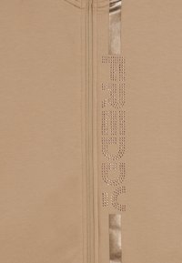 Tessuto beige con una cerniera verticale e borchie metalliche che formano la parola "TRUSSARDI" accanto a due righe verticali lucide.