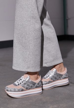 Person iført grå bukser med vide ben og pink og sølvfarvede platåsneakers, der står på en grå gulv indendørs.