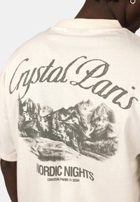Cream cotton t-shirt featuring a graphic design with "Crystal Paris" text and a scenic mountain illustration, labeled "NORDIC NIGHTS."