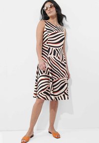 Ärmelloses Kleid mit einem wirbelnden Muster in Schwarz, Weiß und Rot; aus weichem Stoff; knielang; kombiniert mit orangen Pantoletten.