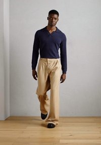 Camisa polo de manga larga en color navy combinada con pantalones anchos beige y zapatos negros, presentada contra una pared clara y texturizada y un suelo de madera.
