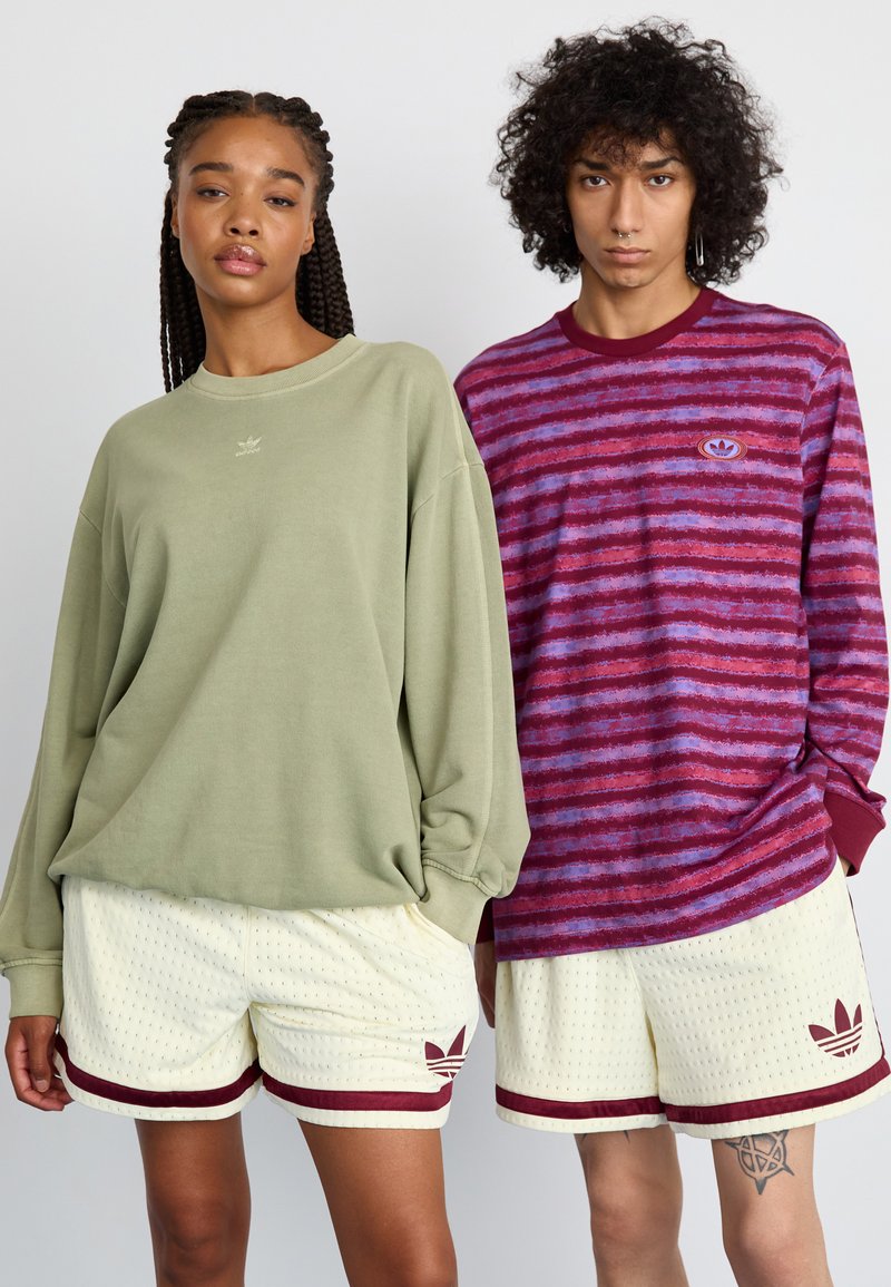 Sweatshirt oversized kaki avec un petit logo, associé à un short en mesh crème avec des rayures maroon et du branding ; le modèle à gauche a des tresses.