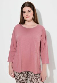 Rosa, leichtes Langarmoberteil mit Rundhalsausschnitt und seitlichen Schlitzen, mit einer Vordertasche; kombiniert mit braunen gemusterten Hosen.