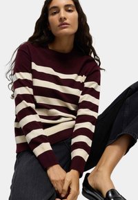 Bordeaux- und cremefarbener gestreifter Pullover mit Rundhalsausschnitt und gerippten Bündchen, kombiniert mit schwarzen Jeans und Mokassins. Die Textur wirkt weich und gestrickt.