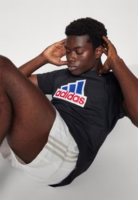 Černé tričko s velkým červeným a modrým logem Adidas, spárované s bílými šortkami s třemi vertikálními šedými pruhy.