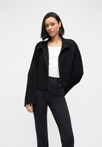 Jachetă neagră scurtă cu fermoar, mâneci largi și două buzunare frontale, purtată peste un top alb și pantaloni negri mulați. Textură netedă a țesăturii.