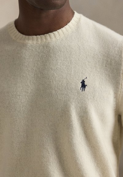 Close-up of a dark-skinned person wearing a cream knit sweater with a small navy polo player logo on the chest.