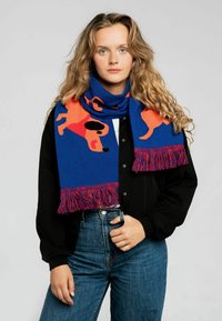 Niebieski szalik z pomarańczowym wzorem psów i frędzlami; wykonany z miękkiego materiału, zarzucony na szyję, podkreślający żywe kolory i zabawny design.