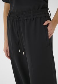 Čierne nohavice s elastickým pásom a bočnými vreckami, s povoľovacím šnúrkou so striebornými koncovkami a hladkou, jemnou textúrou.