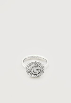 Bague en argent avec un visage rond présentant un "G" en relief au centre, entouré de deux rangées de petits cristaux clairs.