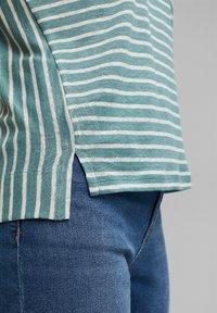 Stribet skjorte i teal og hvid bomuld med sideslids og afslappet pasform, parret med blå denim jeans. Teksturen virker blød.