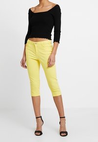 Top negro acanalado sin mangas, con mangas de tres cuartos, y pantalones capri amarillos de corte ajustado. Tacones negros con tiras completan el conjunto.