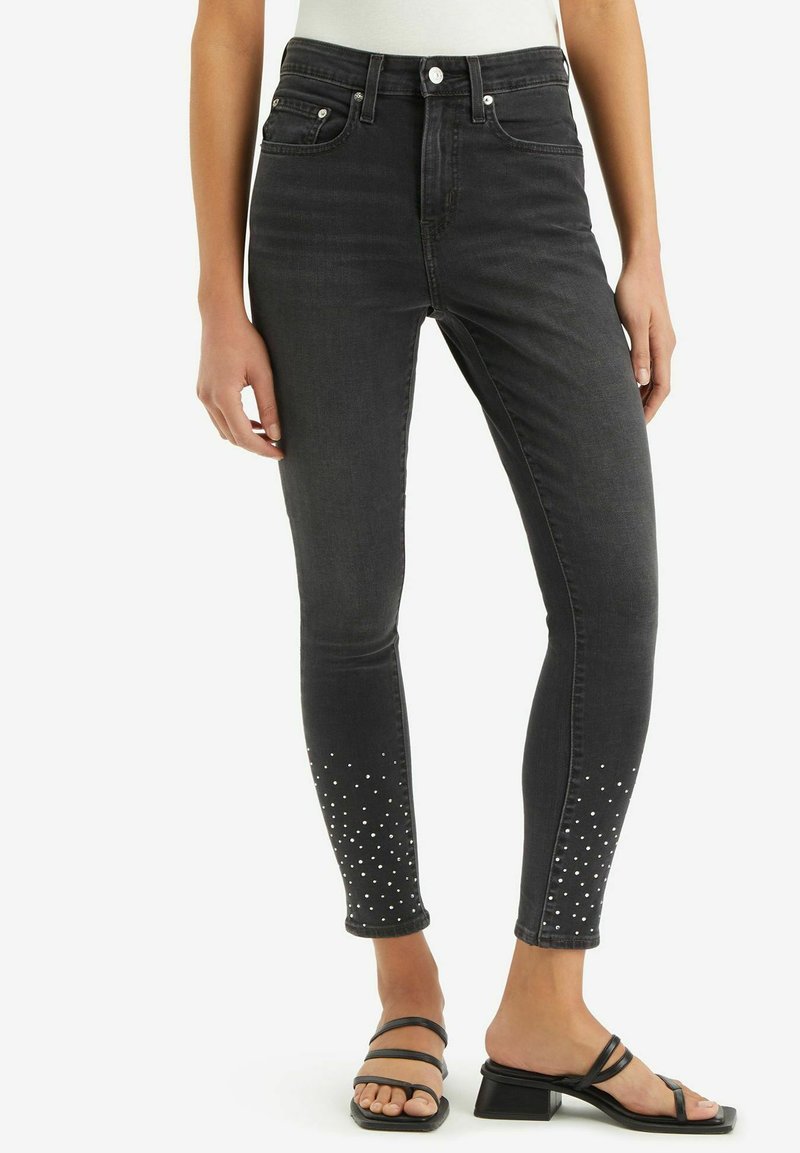 Frau in schwarzen Skinny Jeans mit weißen Nietenverzierungen an den Knöcheln und schwarzen High Heels, stehend vor weißem Hintergrund.