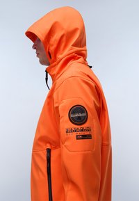 Giacca arancione con cappuccio aderente, tasca laterale con zip e tessuto resistente. Presenta una patch rotonda con logo sulla manica sinistra e dettagli di cucitura.