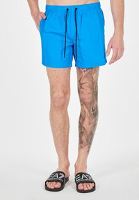 Pantaloni da mare blu con cintura elastica e cordino nero. Abbinati a sandali neri con logo bianco.