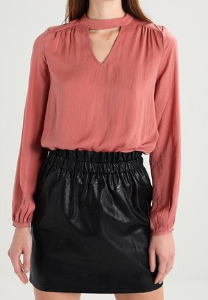Woman wearing a long-sleeve dusty rose blouse with a keyhole neckline and a high-waisted black leather skirt with a gathered waistband.