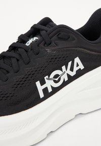 Pantofii sport negri cu partea superioară din plasă, logo alb, guler căptușit și talpă intermediară groasă albă. Prezintă șireturi negre și panouri texturate.