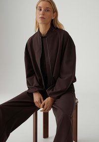 Giacca bomber marrone scuro con colletto alto e cerniera argento, abbinata a pantaloni coordinati marrone scuro. Tessuto liscio e opaco.