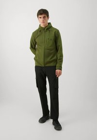 Zelena flis jakna s kapuljačom, patentnim zatvaračem sprijeda i džepom na prsima, u kombinaciji s crnim cargo hlačama sa bočnim džepovima i crnim tenisicama.