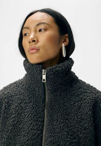 Veste en polaire zippée grise foncée avec un col haut, présentant une surface texturée en boucle et des détails en métal argenté sur la fermeture éclair.