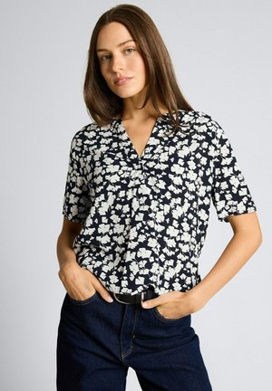 Młoda kobieta z długimi brązowymi włosami ubrana w czarno-białą bluzkę w kwiaty z krótkim rękawem i ciemnoniebieskie dżinsy, ręce w kieszeniach, neutralne tło.