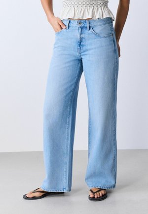Kvinde iført lyseblå jeans med vide ben, hvid cropped top og sorte flipflops, stående med den ene hånd i lommen mod en ensfarvet baggrund.