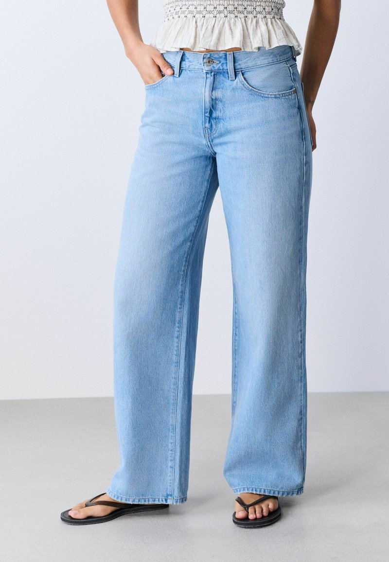 Femme portant un jean large bleu clair, un top court blanc et des tongs noires, debout avec une main dans la poche sur fond uni.