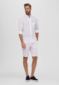 Camisa blanca de lino con mangas remangadas y pequeño bolsillo en el pecho, combinada con pantalones cortos rosa claro y zapatillas negras. Diseño sencillo y ajuste casual.