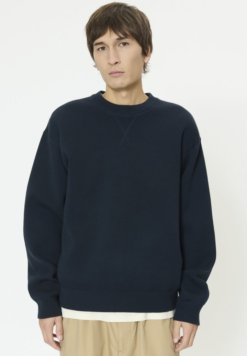 Sweat-shirt bleu marine en matière douce, avec un col rond, des épaules tombantes, des poignets et un ourlet côtelés. Porté par-dessus une chemise claire.