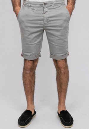 Homme portant un short gris clair retroussé, les mains dans les poches et des chaussures noires sans lacets, debout devant un fond uni.