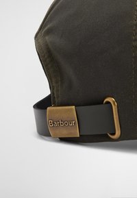 Boné de algodão encerado verde escuro com uma alça ajustável preta. Apresenta uma fivela em latão com "Barbour" gravado. Costuras visíveis.